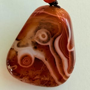 May include: Polished agate pendant with a swirling pattern of brown, white, and reddish-orange hues. The stone is teardrop-shaped and strung on a black cord with a red bead and gold-colored accent.