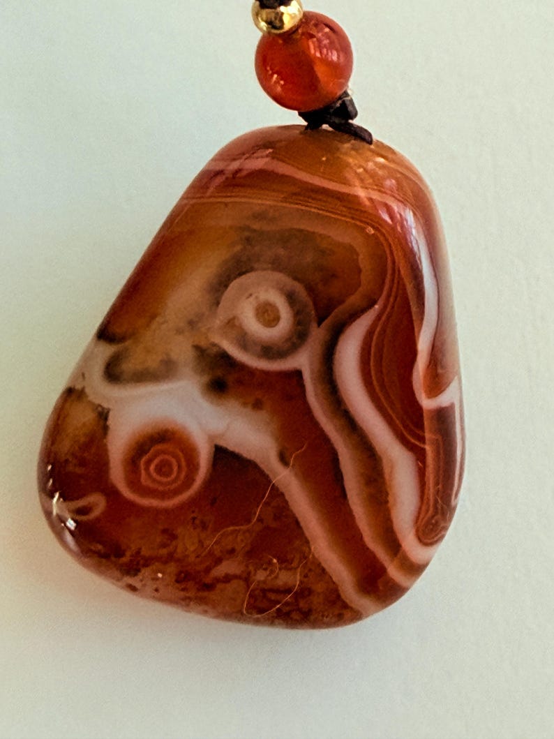 May include: Polished agate pendant with a swirling pattern of brown, white, and reddish-orange hues. The stone is teardrop-shaped and strung on a black cord with a red bead and gold-colored accent.