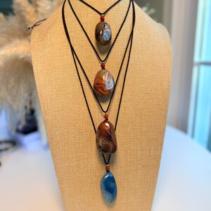 May include: Four gemstone pendant necklaces displayed on a tan mannequin. The necklaces feature polished, oval-shaped stones in shades of brown, red, and blue, suspended from black cords. Each necklace has a small, red bead accent.