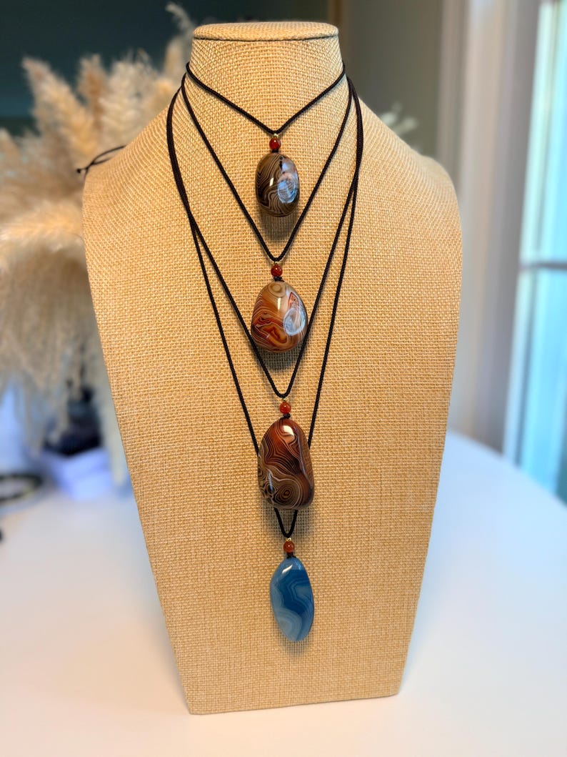 May include: Four gemstone pendant necklaces displayed on a tan mannequin. The necklaces feature polished, oval-shaped stones in shades of brown, red, and blue, suspended from black cords. Each necklace has a small, red bead accent.