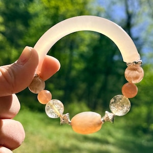 Puede incluir: Una pulsera con una sección translúcida, curva y de color claro y una cadena de cuentas naranjas y transparentes. Las cuentas están conectadas por detalles metálicos plateados. El fondo es una vista borrosa de follaje verde.