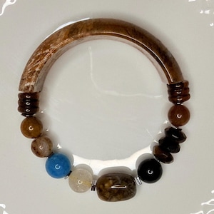 Puede incluir: Una pulsera circular que presenta un gran segmento curvo con patrón marrón, acentuado con varias cuentas que incluyen espaciadores estriados de color marrón oscuro, esferas marrones lisas, una cuenta azul brillante, una cuenta blanca translúcida, una cuenta rectangular marrón y una esfera negra.
