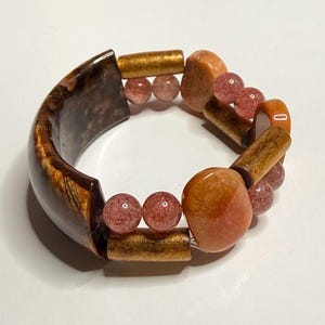 Puede incluir: Una pulsera de múltiples hilos que presenta un segmento grande y curvo de ojo de tigre marrón oscuro, intercalado con cuentas cilíndricas de metal dorado, piezas de piedra lisa en tonos naranja y pequeñas cuentas redondas de color rosa translúcido.