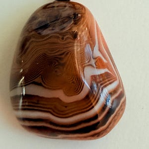 May include: A polished, oval-shaped pendant necklace featuring a banded agate stone. The stone displays layers of brown, white, and black hues. The pendant is suspended from a black cord with a small, red-orange bead.
