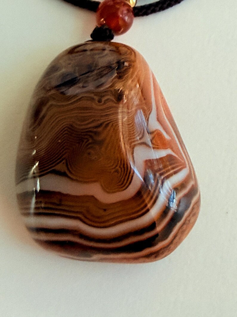 May include: A polished, oval-shaped pendant necklace featuring a banded agate stone. The stone displays layers of brown, white, and black hues. The pendant is suspended from a black cord with a small, red-orange bead.