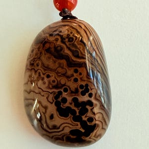 May include: A polished, oval-shaped pendant with a unique pattern of brown and black swirls and spots. The pendant is strung on a dark cord, with a red bead and a gold-colored bead at the top.