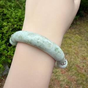 K&ouml;nnte beinhalten: Ein hellgr&uuml;nes Jade-Armreif-Armband. Das Armband hat ein glattes, abgerundetes Design mit subtilen Farbvariationen. Kleine, dekorative silberne Akzente befinden sich an jedem Ende. Der Hintergrund zeigt verschwommenes gr&uuml;nes Laub und Gras.