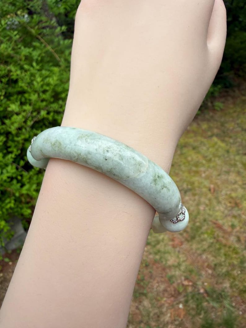 K&ouml;nnte beinhalten: Ein hellgr&uuml;nes Jade-Armreif-Armband. Das Armband hat ein glattes, abgerundetes Design mit subtilen Farbvariationen. Kleine, dekorative silberne Akzente befinden sich an jedem Ende. Der Hintergrund zeigt verschwommenes gr&uuml;nes Laub und Gras.