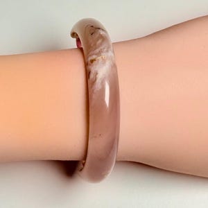 Pu&ograve; includere: Un braccialetto a cerchio traslucido color malva con un motivo marmorizzato bianco sporco. Il braccialetto &egrave; liscio e arrotondato, con una finitura lucida. Il gioiello &egrave; indossato al polso.