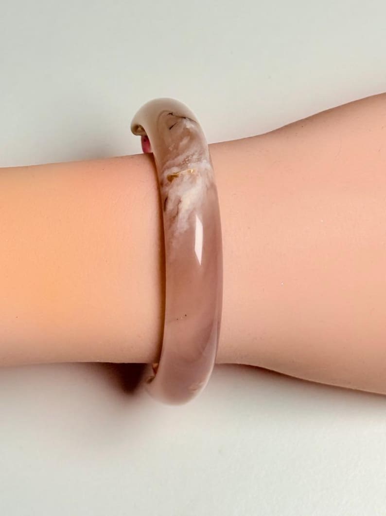 Pu&ograve; includere: Un braccialetto a cerchio traslucido color malva con un motivo marmorizzato bianco sporco. Il braccialetto &egrave; liscio e arrotondato, con una finitura lucida. Il gioiello &egrave; indossato al polso.