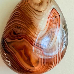 May include: Polished agate pendant with a natural, layered pattern in shades of brown, tan, and white. The stone has a smooth, rounded shape and a glossy finish. A dark brown cord is attached at the top for wearing as a necklace.