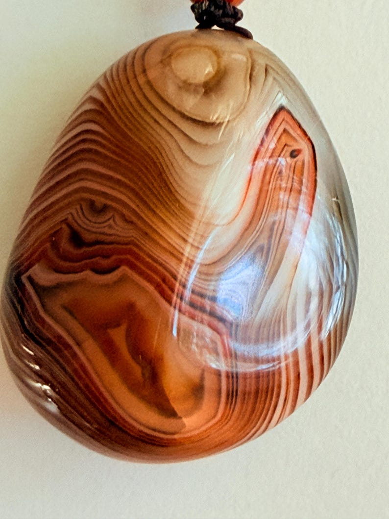 May include: Polished agate pendant with a natural, layered pattern in shades of brown, tan, and white. The stone has a smooth, rounded shape and a glossy finish. A dark brown cord is attached at the top for wearing as a necklace.