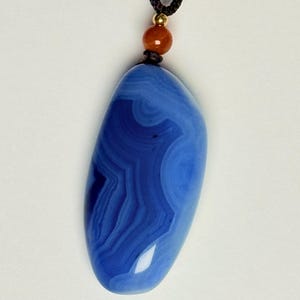 May include: A blue agate pendant with a swirling pattern, suspended from a black cord. The gemstone is a deep shade of blue with lighter blue and white bands. A small orange bead and gold-colored accent are at the top.