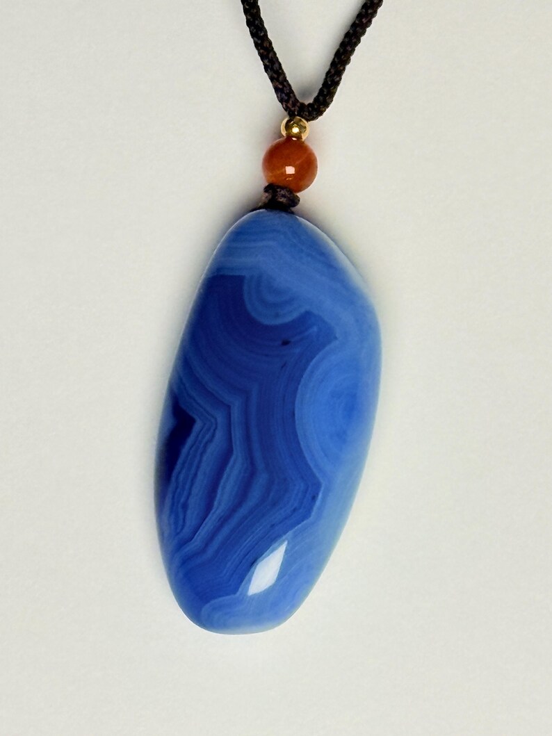 May include: A blue agate pendant with a swirling pattern, suspended from a black cord. The gemstone is a deep shade of blue with lighter blue and white bands. A small orange bead and gold-colored accent are at the top.