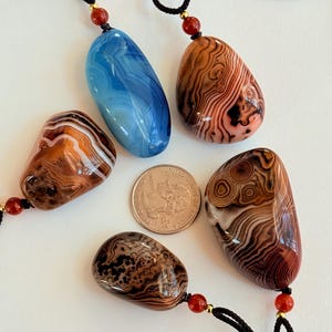 May include: Five polished stone pendants with black cords and red and gold accents. The stones are various colors, including blue, brown, and orange, and have unique patterns. A quarter is included for size reference.