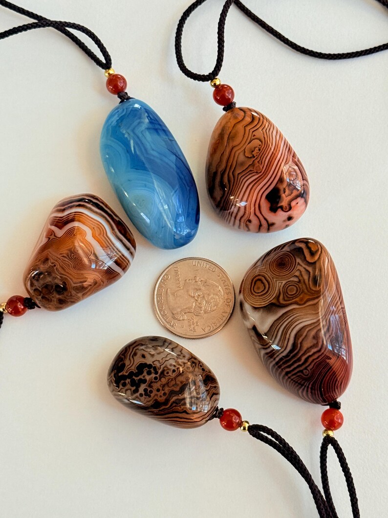 May include: Five polished stone pendants with black cords and red and gold accents. The stones are various colors, including blue, brown, and orange, and have unique patterns. A quarter is included for size reference.