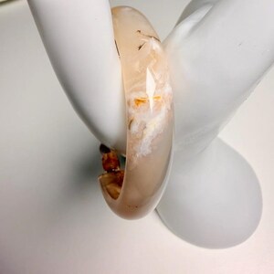Pu&ograve; includere: Un braccialetto a cerchio traslucido beige con inclusioni bianche e arancioni. Il braccialetto &egrave; liscio e arrotondato, con una finitura lucida. Il braccialetto &egrave; esposto su una mano di manichino bianca.