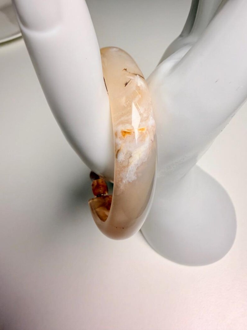 Pu&ograve; includere: Un braccialetto a cerchio traslucido beige con inclusioni bianche e arancioni. Il braccialetto &egrave; liscio e arrotondato, con una finitura lucida. Il braccialetto &egrave; esposto su una mano di manichino bianca.