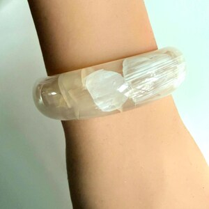 Puede incluir: Una pulsera tipo brazalete ancha y translúcida con inclusiones blancas opacas similares a cristales incrustadas. La pulsera tiene una superficie lisa, pulida y redondeada, con un aspecto transparente similar a la resina que deja pasar la luz.
