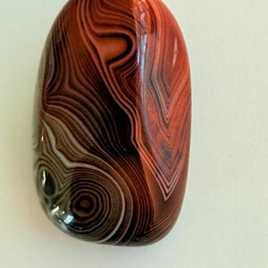 May include: A polished, oval-shaped pendant with a swirling pattern of brown, black, and white bands. The pendant is predominantly reddish-brown and has a glossy finish. A dark brown cord is attached at the top.