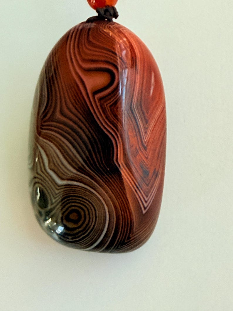May include: A polished, oval-shaped pendant with a swirling pattern of brown, black, and white bands. The pendant is predominantly reddish-brown and has a glossy finish. A dark brown cord is attached at the top.
