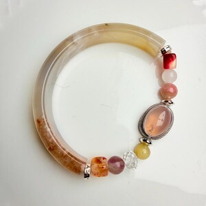 Pu&ograve; includere: Un braccialetto di pietre preziose con una fascia grande marrone chiaro e crema e una variet&agrave; di perline colorate. Le perline includono pietre rosa, rosse, arancioni e trasparenti e una grande pietra rosa ovale.