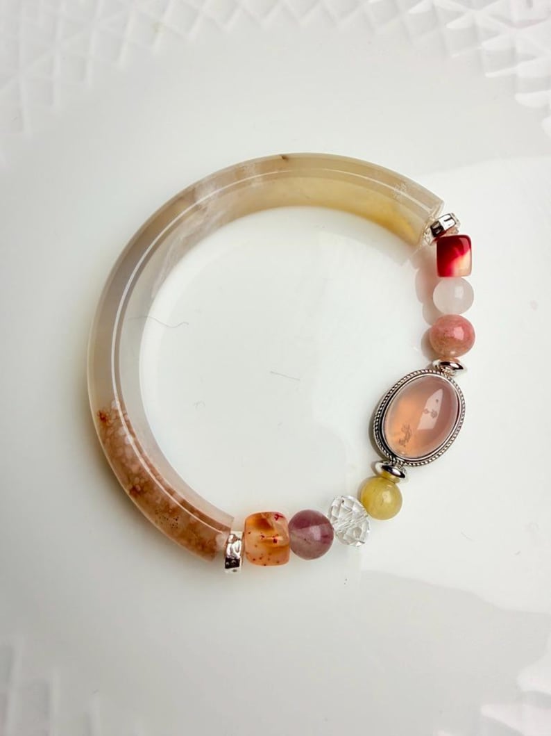 Pu&ograve; includere: Un braccialetto di pietre preziose con una fascia grande marrone chiaro e crema e una variet&agrave; di perline colorate. Le perline includono pietre rosa, rosse, arancioni e trasparenti e una grande pietra rosa ovale.