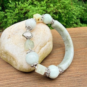K&ouml;nnte beinhalten: Ein hellgr&uuml;nes Jade-Armband mit silbernen Akzenten. Das Armband besteht aus runden und quadratischen Jadeperlen sowie einem gebogenen Jade-Armreif. Das Armband wird auf einem hellen Stein vor einem Holzuntergrund pr&auml;sentiert.