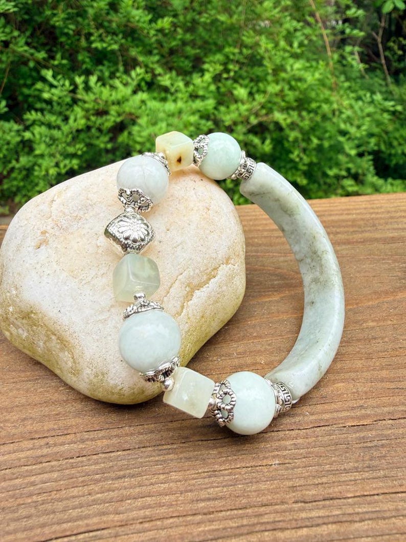 K&ouml;nnte beinhalten: Ein hellgr&uuml;nes Jade-Armband mit silbernen Akzenten. Das Armband besteht aus runden und quadratischen Jadeperlen sowie einem gebogenen Jade-Armreif. Das Armband wird auf einem hellen Stein vor einem Holzuntergrund pr&auml;sentiert.