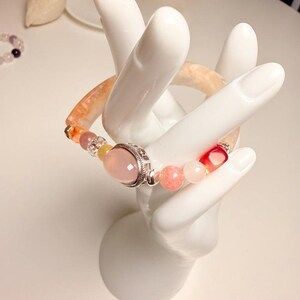 Pu&ograve; includere: Un braccialetto di perline con una grande gemma rosa incastonata in metallo color argento. Il braccialetto include una variet&agrave; di perline colorate nelle tonalit&agrave; del rosa, dell'arancione e del trasparente. Il braccialetto &egrave; esposto su un portagioielli bianco a forma di mano.