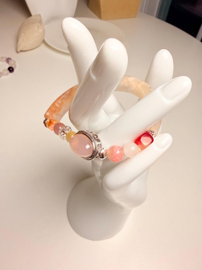 Pu&ograve; includere: Un braccialetto di perline con una grande gemma rosa incastonata in metallo color argento. Il braccialetto include una variet&agrave; di perline colorate nelle tonalit&agrave; del rosa, dell'arancione e del trasparente. Il braccialetto &egrave; esposto su un portagioielli bianco a forma di mano.