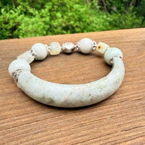 K&ouml;nnte beinhalten: Ein Jade-Armband mit einem gebogenen, hellgr&uuml;nen Mittelst&uuml;ck und verschiedenen wei&szlig;en und hellgr&uuml;nen Perlen. Das Armband hat silberfarbene Akzente und wird auf einer Holzoberfl&auml;che pr&auml;sentiert.
