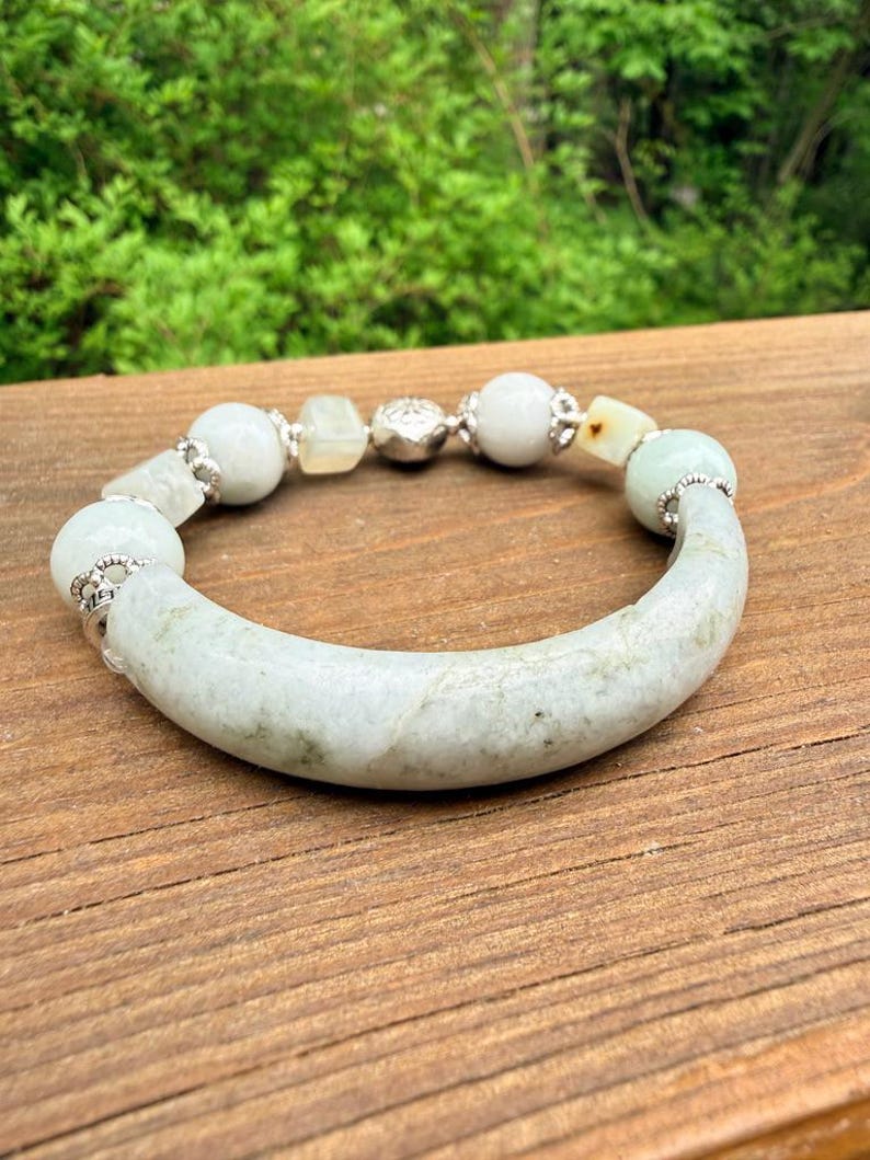 K&ouml;nnte beinhalten: Ein Jade-Armband mit einem gebogenen, hellgr&uuml;nen Mittelst&uuml;ck und verschiedenen wei&szlig;en und hellgr&uuml;nen Perlen. Das Armband hat silberfarbene Akzente und wird auf einer Holzoberfl&auml;che pr&auml;sentiert.