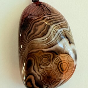 May include: A polished, oval-shaped pendant with a swirling pattern of brown, tan, and white bands. The pendant has a glossy finish and is strung on a black cord with a red bead and gold-colored bead.