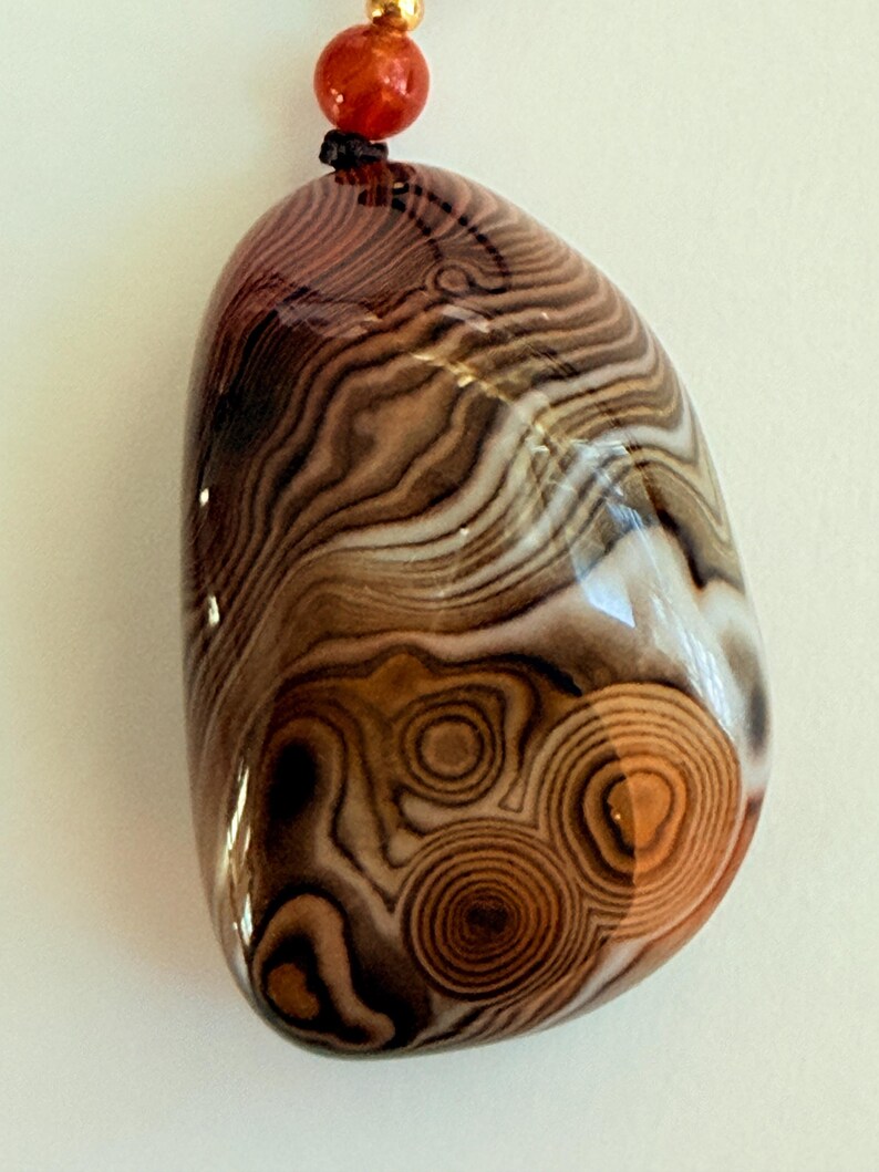 May include: A polished, oval-shaped pendant with a swirling pattern of brown, tan, and white bands. The pendant has a glossy finish and is strung on a black cord with a red bead and gold-colored bead.