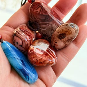 May include: A collection of polished agate pendants in various shapes and colors, including red, brown, white, and blue. Each stone features unique swirling patterns and is strung on a cord with a bead.