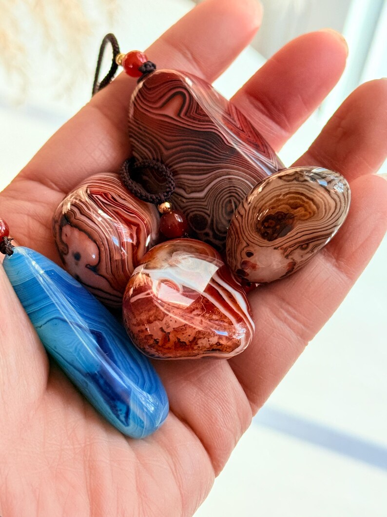 May include: A collection of polished agate pendants in various shapes and colors, including red, brown, white, and blue. Each stone features unique swirling patterns and is strung on a cord with a bead.