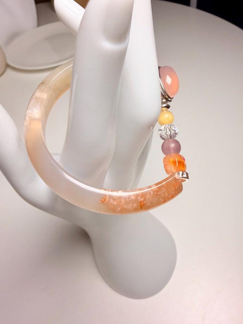 Pu&ograve; includere: Un braccialetto a cerchio color pesca chiaro con una sezione maculata arancione e bianca. &Egrave; adornato da una fila di perline colorate, tra cui una grande pietra rosa e perline gialle, trasparenti e arancioni pi&ugrave; piccole. Il braccialetto &egrave; esposto su una mano bianca.