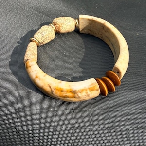May include: A circular bracelet featuring a combination of smooth, curved beige stone segments with brown marbling, two oval-shaped crackled beige beads, and three small, flat, circular brown wooden discs separated by thin gold-toned metal spacers.