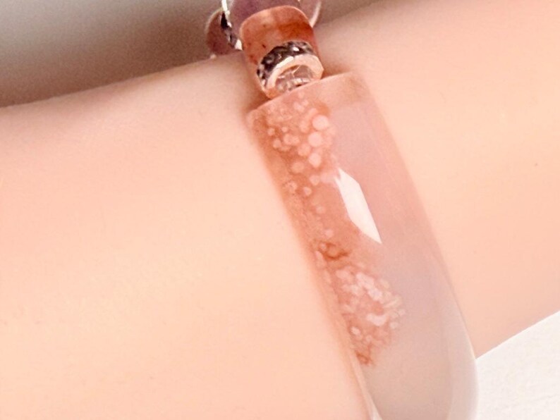 Pu&ograve; includere: Primo piano di un braccialetto con una grande perla rettangolare traslucida con una tonalit&agrave; rosa chiaro e un motivo di macchie marrone rossiccio. Il braccialetto ha un connettore in metallo color argento e perline rotonde pi&ugrave; piccole.