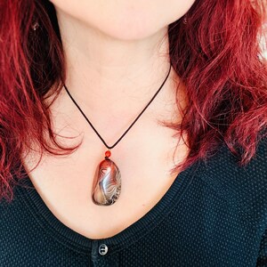 May include: A pendant necklace featuring a large, polished stone with swirling patterns in shades of brown, cream, and black. The stone is suspended from a black cord with a small red bead accent. The necklace is a unique piece of jewelry.