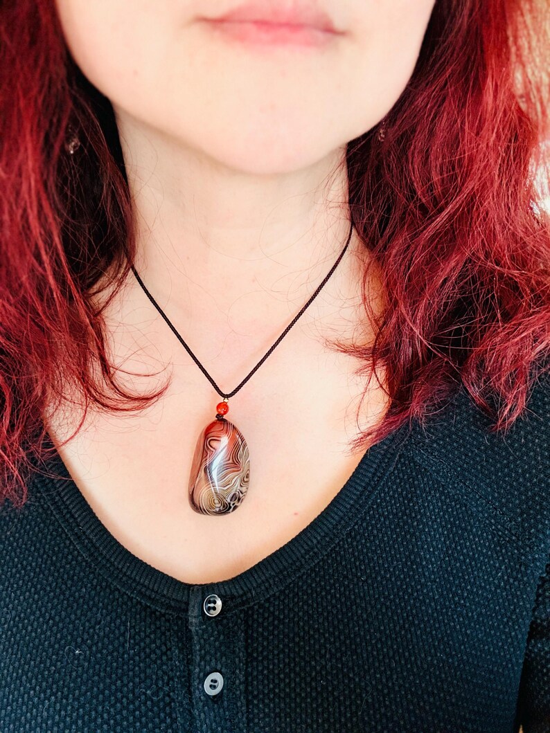 May include: A pendant necklace featuring a large, polished stone with swirling patterns in shades of brown, cream, and black. The stone is suspended from a black cord with a small red bead accent. The necklace is a unique piece of jewelry.