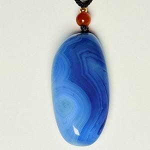 May include: A blue agate pendant necklace with a black cord. The pendant is an oval shape with concentric bands of light and dark blue. A small red bead and gold-colored bead accent the necklace.