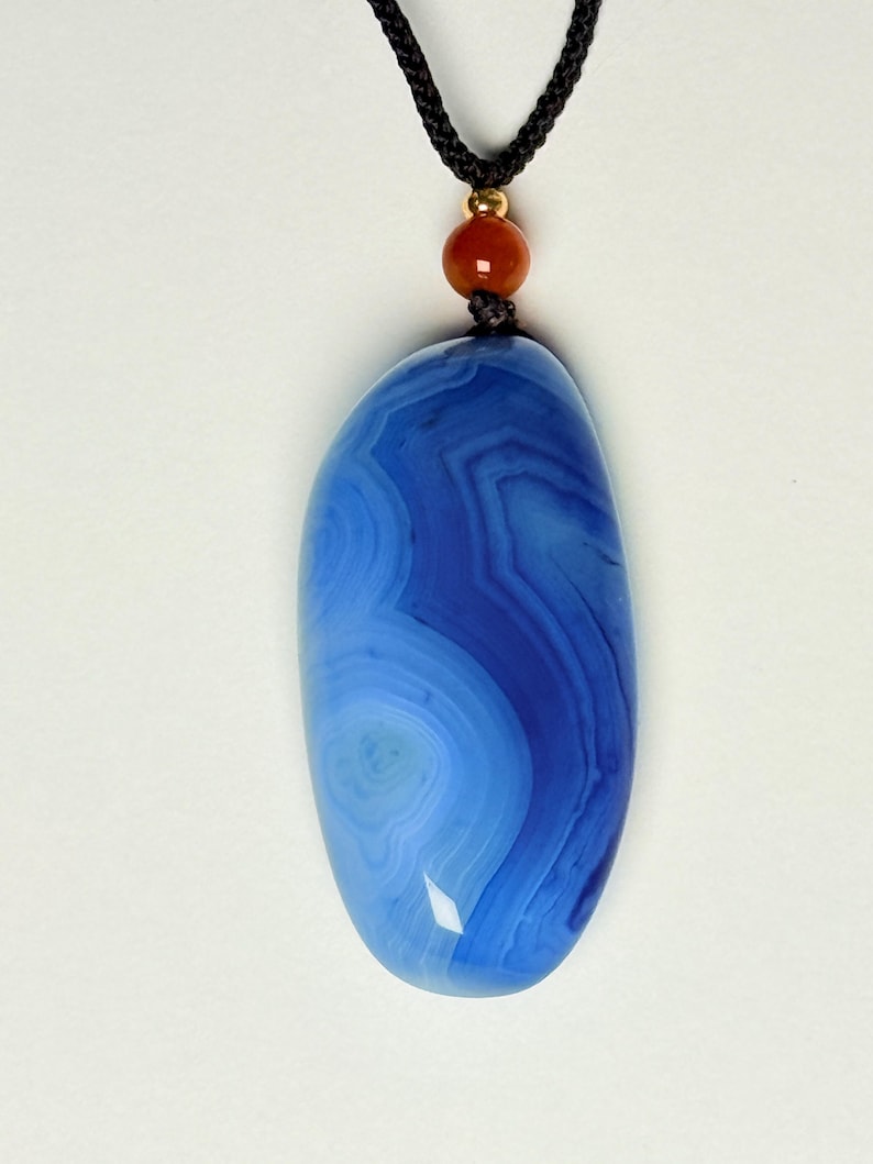 May include: A blue agate pendant necklace with a black cord. The pendant is an oval shape with concentric bands of light and dark blue. A small red bead and gold-colored bead accent the necklace.
