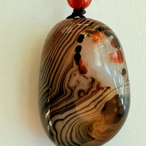 May include: A polished, oval-shaped pendant with a brown and cream striped pattern. The pendant has a glossy finish and is strung on a black cord with a red bead and gold-colored bead at the top. The stone has a natural, organic appearance.