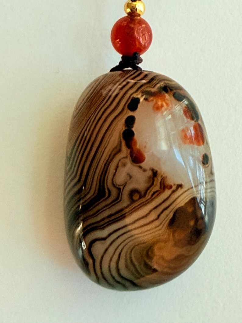 May include: A polished, oval-shaped pendant with a brown and cream striped pattern. The pendant has a glossy finish and is strung on a black cord with a red bead and gold-colored bead at the top. The stone has a natural, organic appearance.