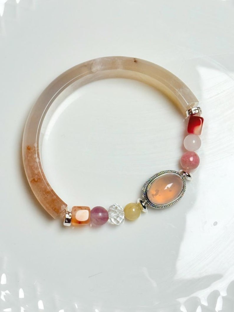 Pu&ograve; includere: Un braccialetto con una grande fascia circolare marrone chiaro e una variet&agrave; di perline colorate. Le perline includono pietre rosa, rosse, gialle e trasparenti e una grande pietra rosa ovale in un'incastonatura d'argento.
