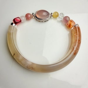 Pu&ograve; includere: Un braccialetto di pietre preziose con una grande pietra rosa ovale incastonata in una cornice d'argento e una variet&agrave; di perline colorate. Il braccialetto include perline rosa, gialle e trasparenti e due sezioni curve marrone chiaro.
