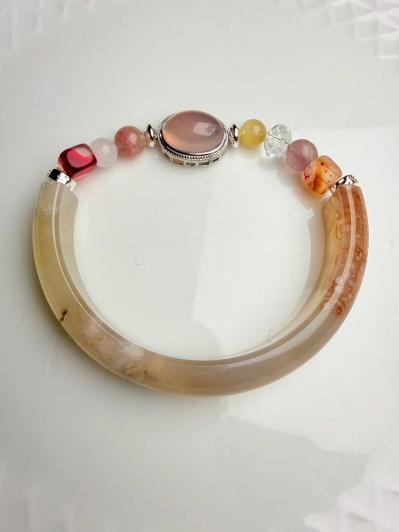 Pu&ograve; includere: Un braccialetto di pietre preziose con una grande pietra rosa ovale incastonata in una cornice d'argento e una variet&agrave; di perline colorate. Il braccialetto include perline rosa, gialle e trasparenti e due sezioni curve marrone chiaro.