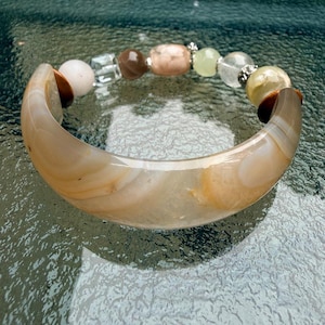 Puede incluir: Una pulsera que presenta un gran segmento de ágata beige translúcido y curvado, conectado a una hilera de cuentas variadas. Las cuentas incluyen esferas blancas, transparentes, marrones, color melocotón, verde pálido y metálicas con espaciadores plateados.